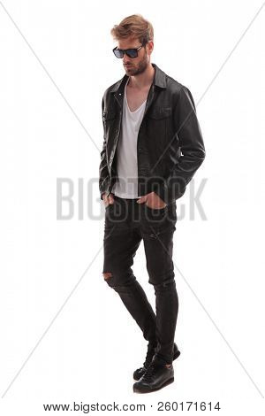 relaxed fashion man with sunglasses and black leather jacket walking on white background and looking down to side, full body picture