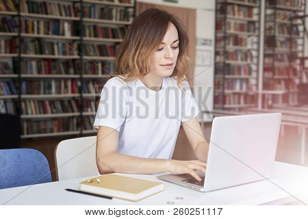 Young Woman Long Hair Analyst Working At Co-working Office On Laptop, Bookshelf Behind. Businesswoma