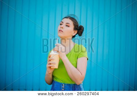 Portrait Of A Girl Against A Bright Blue Wall. Emotional Girl Holding A Plastic Glass With A Refresh