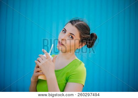 Portrait Of A Girl Against A Bright Blue Wall. Emotional Girl Holding A Plastic Glass With A Refresh
