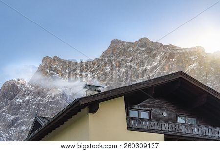 Cozy Winter Scene With The Rooftop Of A House, A Smoky Chimney, And The Snow-capped Alps Mountains, 