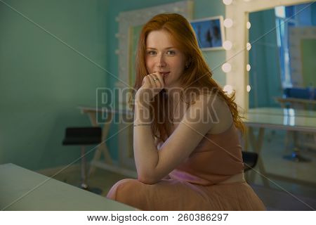 Portrait In Warm Tones, An Attractive Young Woman With Long Red Hair Sitting On A Chair In The Dress