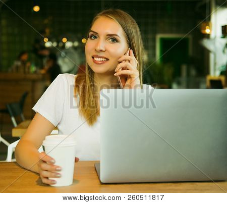 Young beautiful blond girl sitting in a coffee shop and working on computer. Image of happy woman using laptop and smartphone in cafe.