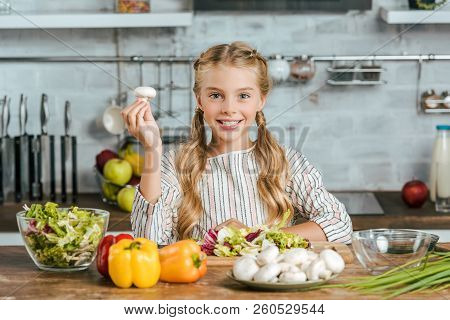 Beautiful Little Child With Mushroom And Various Raw Vegetables Looking At Camera While Making Salad