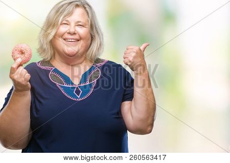 Senior plus size caucasian woman eating sugar donut over isolated background pointing and showing with thumb up to the side with happy face smiling