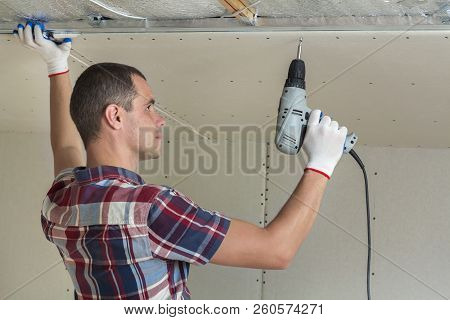 Young Man In Usual Clothing And Work Gloves Fixing Drywall Suspended Ceiling To Metal Frame Using El