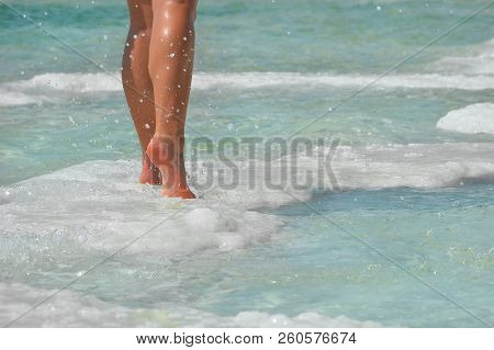 Female Feet With Manicure On Stone, Covered With Salt Formations Between The Waves In The Water. Cry