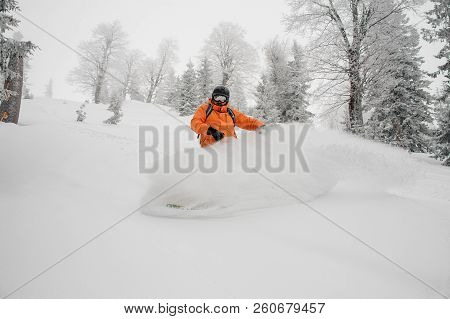 Male Snowboarder Dressed In Orange Sportswear Riding Down The Powder Mountain Hill On Cloudy Day. Sn