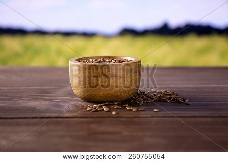 Lot Of Whole Fresh Brown Flaxseeds With Wooden Bowl With Green Wheat Field In Background