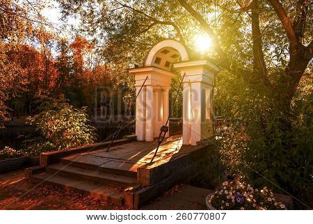 Ancient White Arch Of The Entrance To The Bridge In The City Park, In Autumn The Sun's Rays Make The