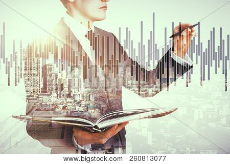 Handsome European Busnessman Holding Book And Drawing Forex Chart On Abstract City Background. Educa