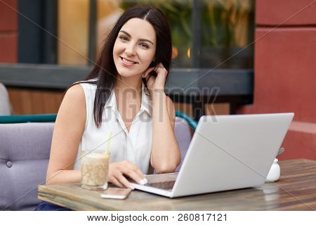 Satisfied Pleasant Looking Freelancer Looks Distantly With Notebook, Drinks Milkshake, Looks Positiv