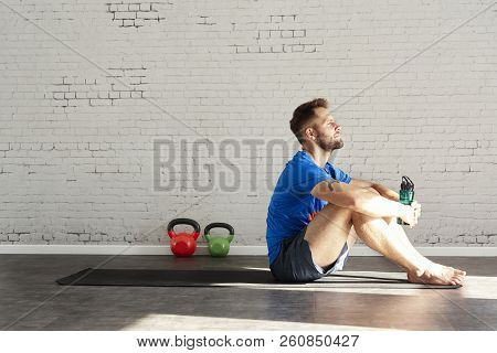 Muscular Athlete Sitting In Sunny Sport Club, Relaxed After Workout, Bottle Of Water In Hands. Space