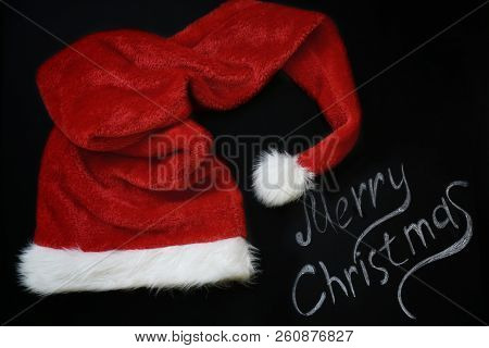 Red Christmas Cap Santa Claus On Chalk, Slate With Chalk Painted Inscription Merry Christmas