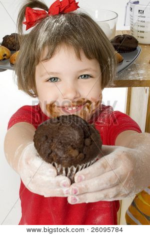 Bella ragazza quattro anni consegnando muffin al cioccolato chip alla macchina fotografica.  Cottura ingredienti in backgro