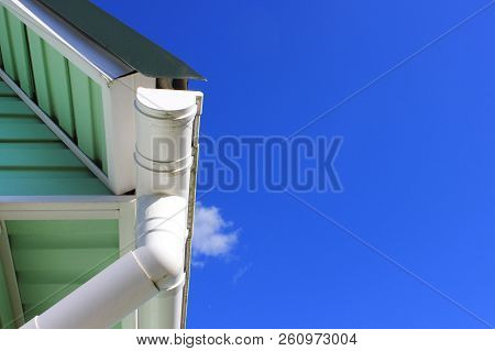 House Corner With Gutter, Roof Top Tiles, Drainage System And Azure Wall Detail On Empty Blue Sky Ba