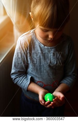 Little Todder Girl Stanting Next To The Window And Holding Green Polka Dotted Easter Egg In Her Palm