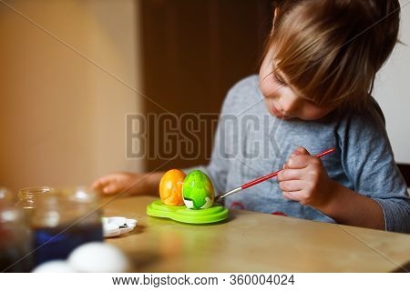 Todder Girl Sitting At The Table And Painting Easter Eggs, Holding Brush In Her Hand