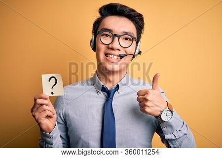 Young handsome chinese call center agent man holding paper with question mark symbol happy with big smile doing ok sign, thumb up with fingers, excellent sign