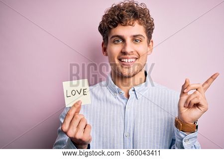 Young blond romantic man with curly hair holdiing paper with love message for valentines day very happy pointing with hand and finger to the side