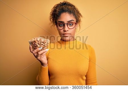 Young african american afro girl holding bowl with healthy peanuts over yellow background with a confident expression on smart face thinking serious