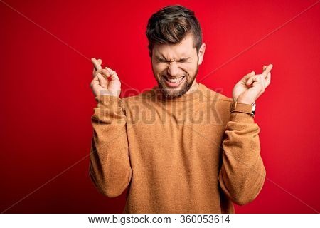 Young blond man with beard and blue eyes wearing casual sweater over red background gesturing finger crossed smiling with hope and eyes closed. Luck and superstitious concept.