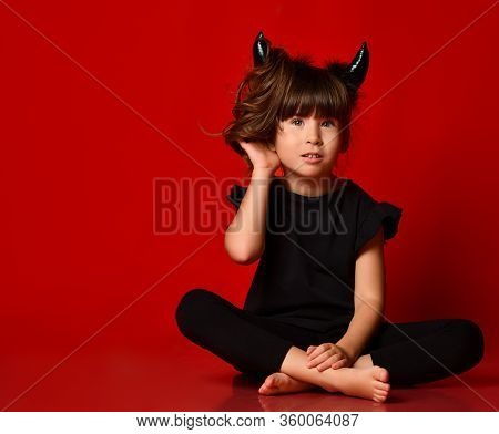 Little Brunette Girl With Devil Horns, Ponytail, In Black Blouse And Leggings, Barefoot. She Is Smil