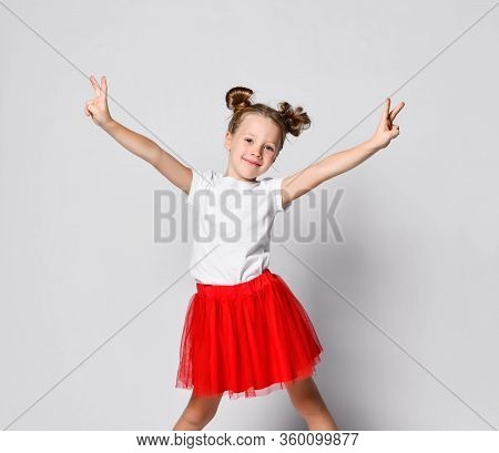 Happy Smiling Blonde Kid Girl In Red Skirt, White T-shirt And Black Sneakers Stands With Her Legs Wi