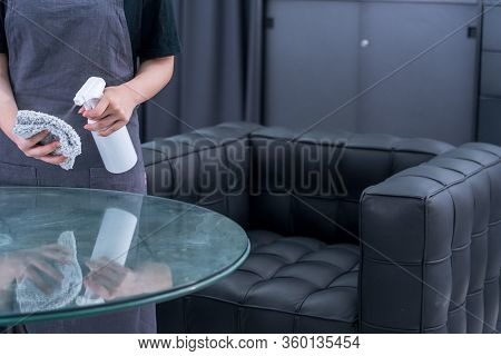 Young Woman Maid In Apron Is Cleaning, Wiping Down Office Glass Table Surface With Spray Bottle Clea