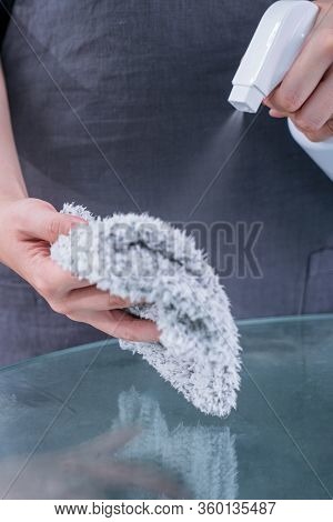 Young Woman Maid In Apron Is Cleaning, Wiping Down Office Glass Table Surface With Spray Bottle Clea
