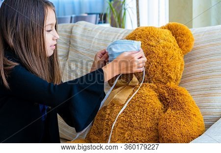 Beautiful Little Girl In A Black Jacket Playing With Her Brown Teddy Bear Who Has Put A Mask To Prot