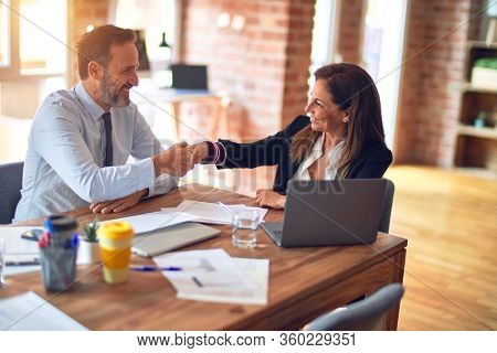 Two middle age business workers smiling happy and confident. Working together with smile on face using laptop doing handsake at the office