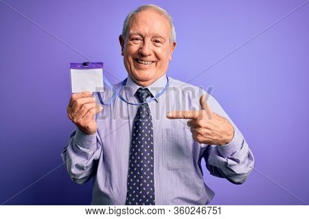 Senior grey haired business man holding identification tag over purple background with surprise face pointing finger to himself