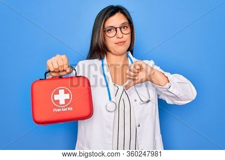 Young doctor woman holding medical first aid kit red box for emergency over blue background with angry face, negative sign showing dislike with thumbs down, rejection concept