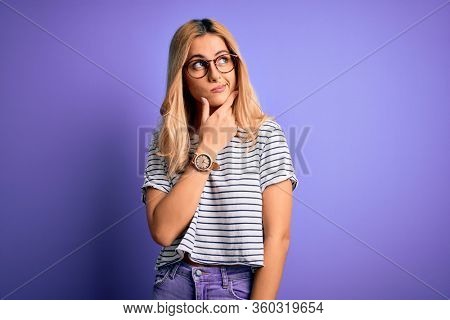 Young beautiful blonde woman wearing striped t-shirt and glasses over purple background Thinking worried about a question, concerned and nervous with hand on chin