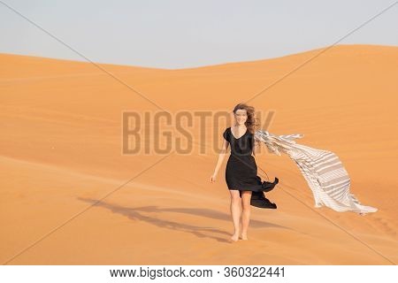A Girl Walks On The Sand In A Deserted Desert. A Woman In A Black Dress With A Cloth In Her Hands In