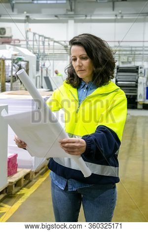 Focused Female Engineer Studying Blueprint Onsite. Middle Aged Woman In Protective Uniform Jacket St