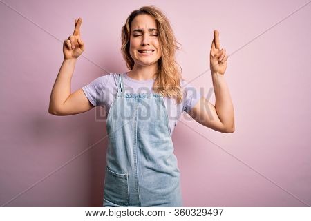 Young beautiful blonde woman wearing casual denim overalls standing over pink background gesturing finger crossed smiling with hope and eyes closed. Luck and superstitious concept.