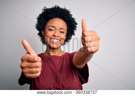 Young beautiful African American afro woman with curly hair wearing casual t-shirt standing approving doing positive gesture with hand, thumbs up smiling and happy for success. Winner gesture.