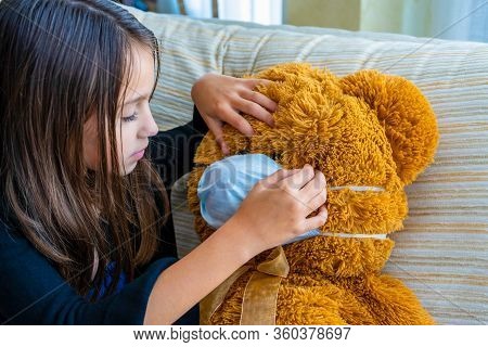 Beautiful Little Girl In A Black Jacket Playing With Her Brown Teddy Bear Who Has Put A Mask To Prot