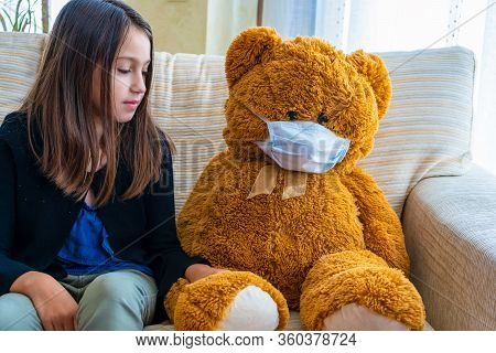 Beautiful Little Girl In A Black Jacket Playing With Her Brown Teddy Bear Who Has Put A Mask To Prot