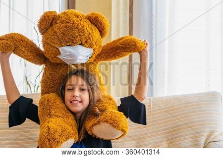 Beautiful Little Girl In A Black Jacket Playing With Her Brown Teddy Bear Who Has Put A Mask To Prot
