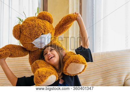 Beautiful Little Girl In A Black Jacket Playing With Her Brown Teddy Bear Who Has Put A Mask To Prot
