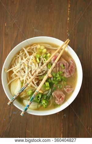Top view of an asian noodle soup with beef, shives, raw onion, bean sprout and chopsticks on top of the white bowl on a wooden background