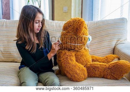 Beautiful Little Girl In A Black Jacket Playing With Her Brown Teddy Bear Who Has Put A Mask To Prot