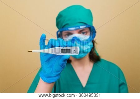 Female Doctor In Green Suit And Blue Mask Gloves And Protective Glasses Withthermometer In Her Hand 