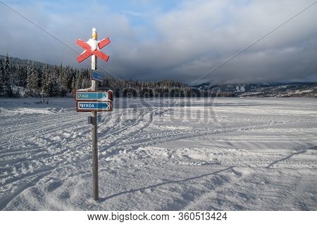 Pole With Directions On A Frozen, Snow-covered Lake With Cloud-covered Mountains And Forests In 
