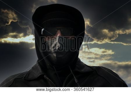 A Man In A Black Protective Mask, Hat, Dark Glasses And A Hood Against A Dramatic Sky. The Concept O