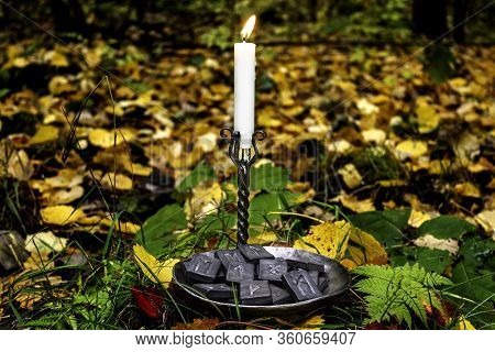 Norse Runes In A Forged Bowl With A Candle On The Background Of Autumn Forest Foliage.