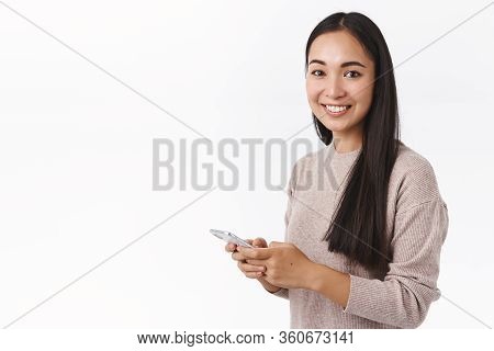 Technology, Young Generation Concept. Attractive Smiling Asian Woman In Sweater, Holding Smartphone 